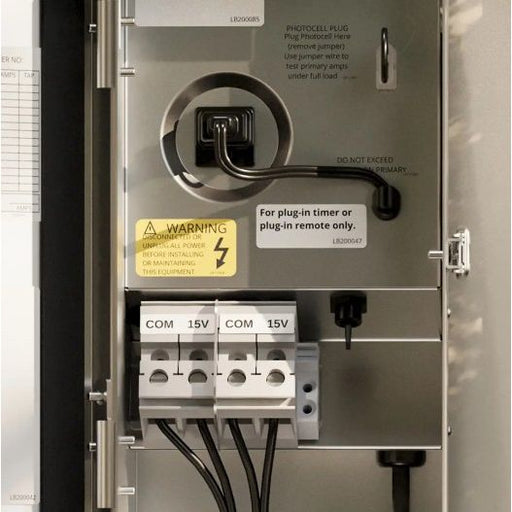 Close-up of an electrical panel door showing a plug-in socket, warning label, terminal connectors labeled COM 15V, and instructional text on a metallic panel.