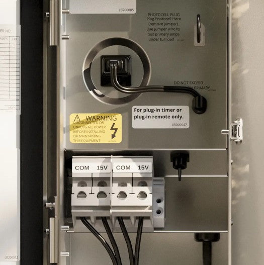 Close-up of an electrical panel door showing a plug-in socket, warning label, terminal connectors labeled COM 15V, and instructional text on a metallic panel.