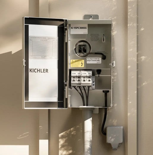 An open Kichler electrical panel box mounted on a beige exterior wall, showing the interior switchgear, terminal connections, and a small attached junction box below.