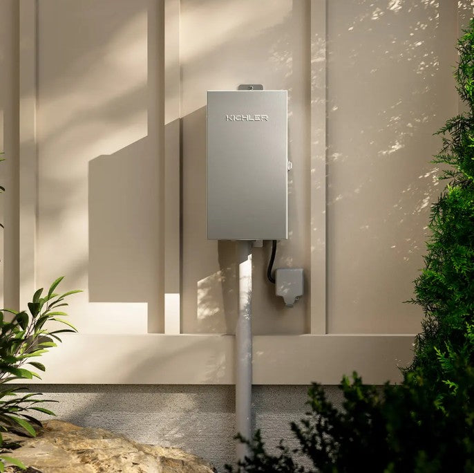 A wall-mounted Rheem water heater in a brushed silver finish, installed outdoors on a beige stucco wall with green plants and natural dappled sunlight.
