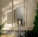 A wall-mounted Rheem water heater in a brushed silver finish, installed outdoors on a beige stucco wall with green plants and natural dappled sunlight.
