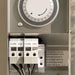Close-up of a mechanical timer switch control panel showing a round dial and terminal connectors labeled COM 15V with wiring visible below.