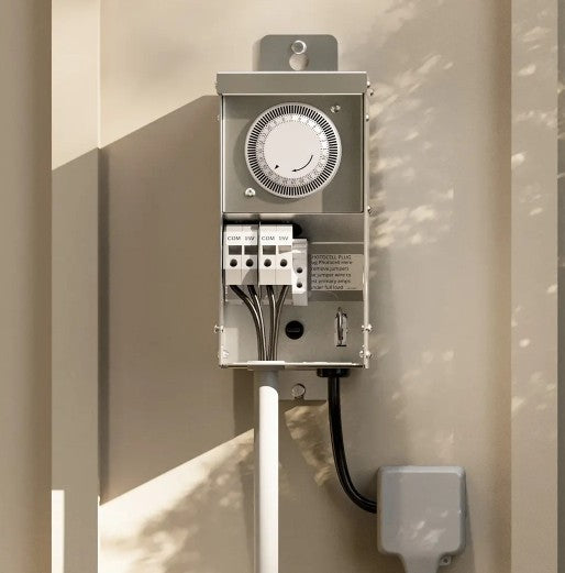 A metal timer electrical switch box with a round dials, mounted on a beige wall with soft leaf shadows.