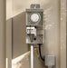 A metal timer electrical switch box with a round dials, mounted on a beige wall with soft leaf shadows.