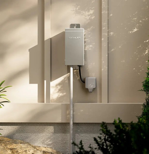 A wall-mounted outdoor electrical unit with a metal enclosure and attached cable, installed on a beige exterior wall with natural shadows and plants nearby.