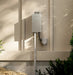 A wall-mounted outdoor electrical unit with a metal enclosure and attached cable, installed on a beige exterior wall with natural shadows and plants nearby.