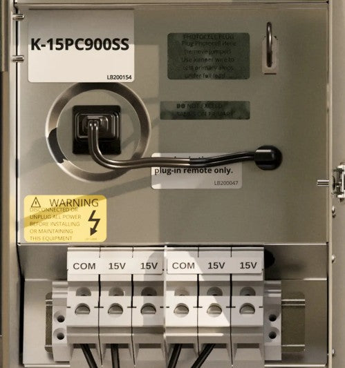 An industrial electrical panel or distribution box labeled K-15PC900SS, with terminal blocks for COM and 15V, warning stickers, and a remote switch handle.