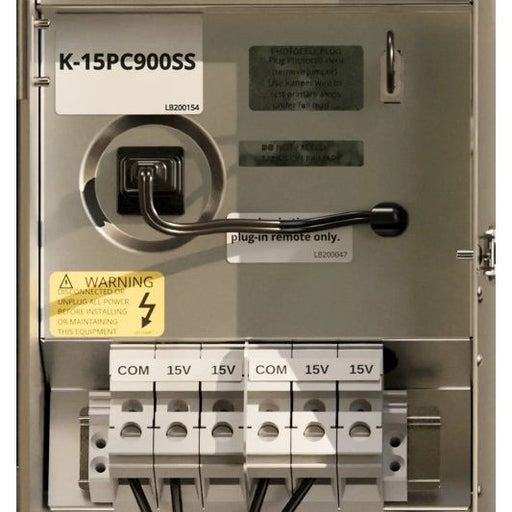 An industrial electrical panel or distribution box labeled K-15PC900SS, with terminal blocks for COM and 15V, warning stickers, and a remote switch handle.
