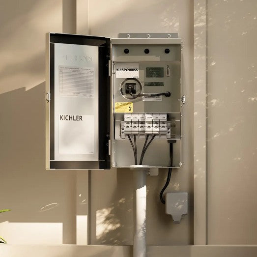 An open Kichler electrical panel mounted on a beige wall, showing the internal terminal blocks, wiring, and switch mechanism. The panel door is open to the left, and the interior metal box is visible against warm sunlight and shadows.