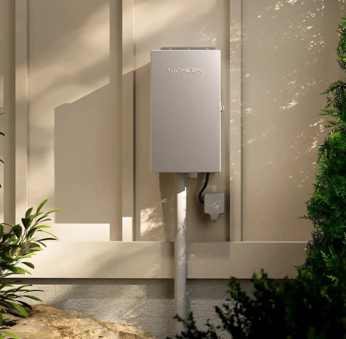A silver NICHILSEN outdoor electrical storage system mounted on a beige stucco wall with soft sunlight and shadows, flanked by green plants.