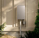A silver NICHILSEN outdoor electrical storage system mounted on a beige stucco wall with soft sunlight and shadows, flanked by green plants.