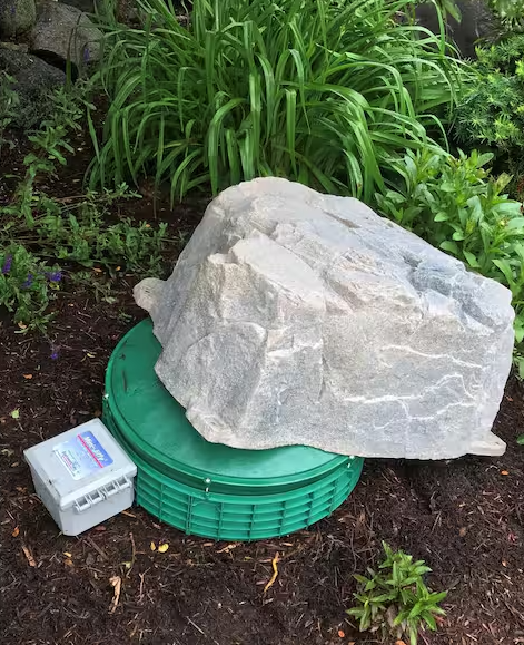 A large natural-looking gray stone or rock placed on top of a round green lid, outdoors in mulch with green plants in the background. A small white control box is also visible beside the setup.
