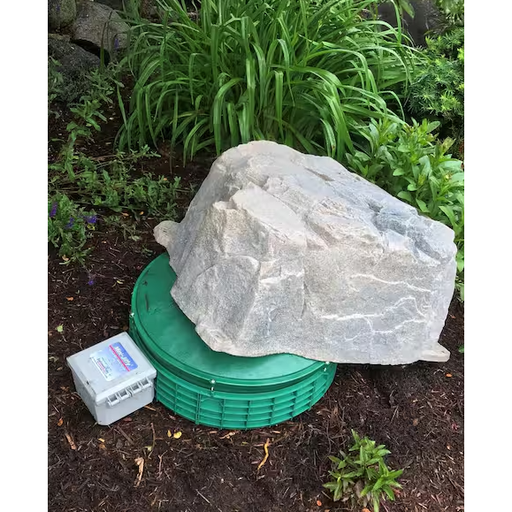A large natural-looking gray stone or rock placed on top of a round green lid, outdoors in mulch with green plants in the background. A small white control box is also visible beside the setup.