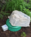 A large natural-looking gray stone or rock placed on top of a round green lid, outdoors in mulch with green plants in the background. A small white control box is also visible beside the setup.