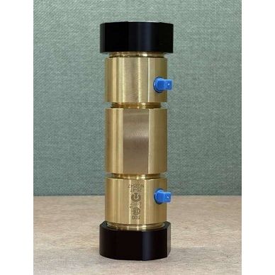 A brass cylindrical component with black end caps and small blue side valves, standing upright on a light surface against a textured green backdrop.